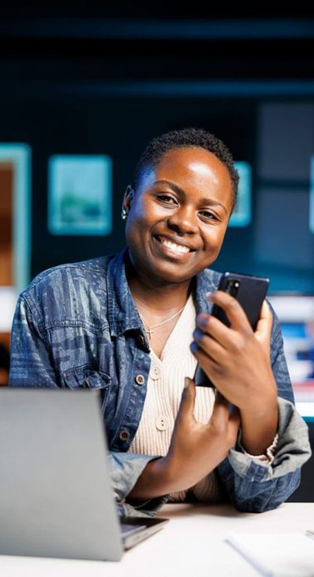 Mulher com expressão feliz em frente a um notebook, olhando para o celular, sentada à mesa