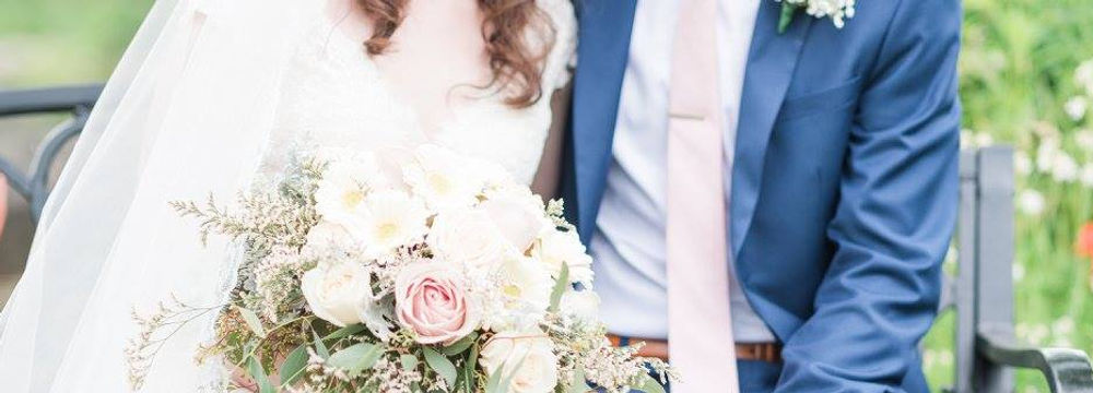 A bride with a long veil and in a lace white dress holds her wedding bouquet and sits on a bench next to a groom in a navy blue suit with a pink tie.