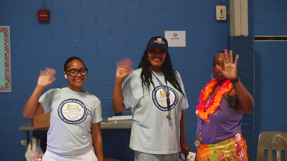 Three smiling women wave in a blue school hall; one sign reads 8th Grade. They wear matching shirts and colorful leis.