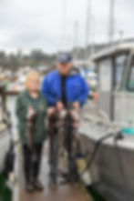 Two people on a dock hold large fish, smiling. They're wearing jackets; boats nearby. Overcast sky and moored boats in the background.