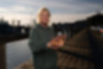 Woman in a green hoodie holds a fish on a wooden pier with boats in the background, under a partly cloudy sky at sunset.