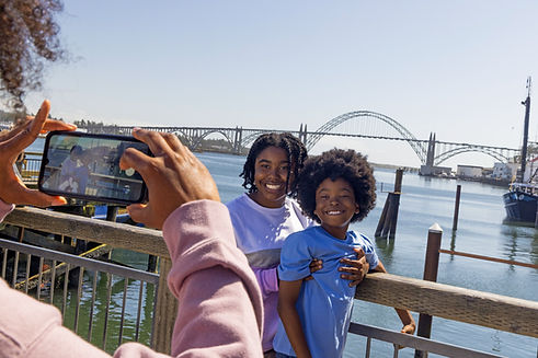 A sister and brother pose on the dock in front of the Yaquina Bay Bridge while some takes a photo on their phone.