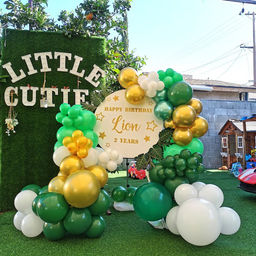 Jungle-themed 2nd birthday setup for Lion with green and gold balloon arch, "Little Cutie" sign on grass backdrop, and outdoor decor.