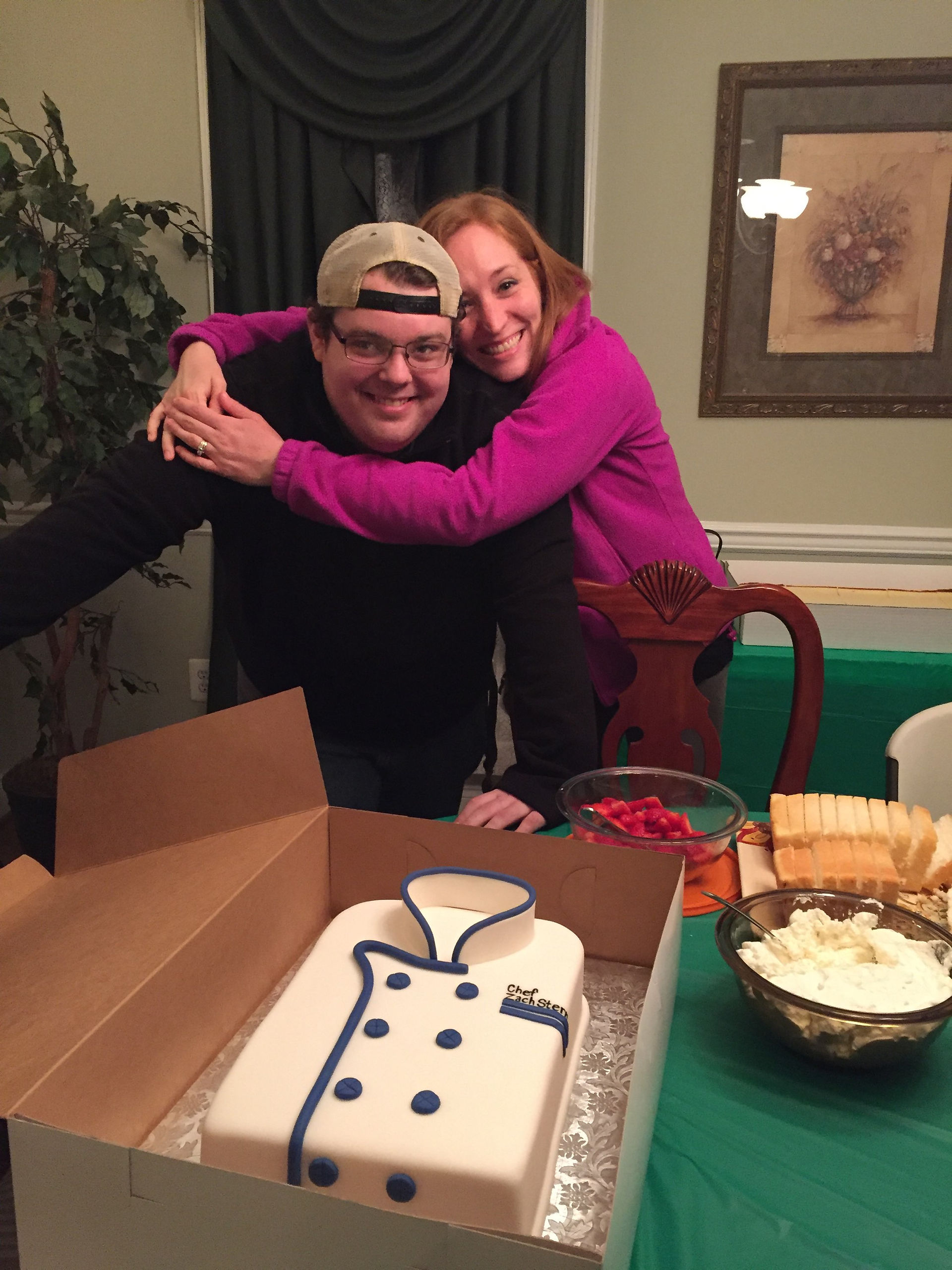 Zach’s aunt hugs him proudly as he stands in front of a chef themed cake celebrating his journey to becoming a chef.