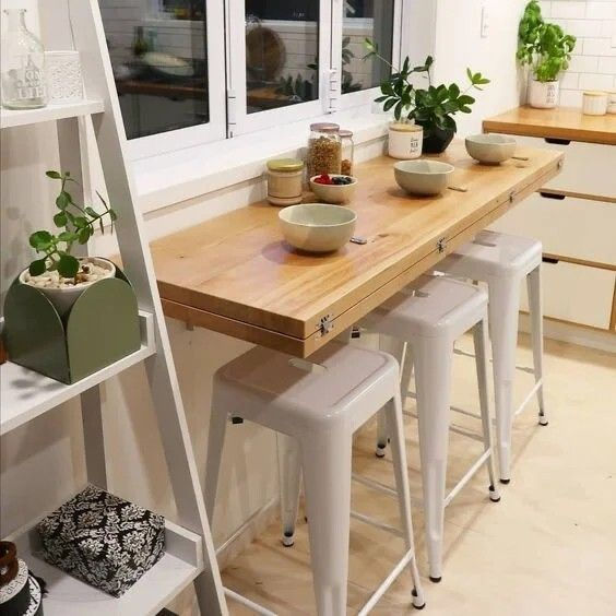 Kitchen with fold down wooden counter, white stools, bowls, and plants. Shelves hold small decor items. Bright, clean, and modern ambiance.