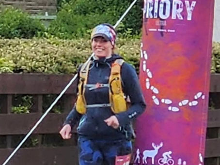 A woman smiling and running at the start of a race. Priory Ultra 100km arch in the background