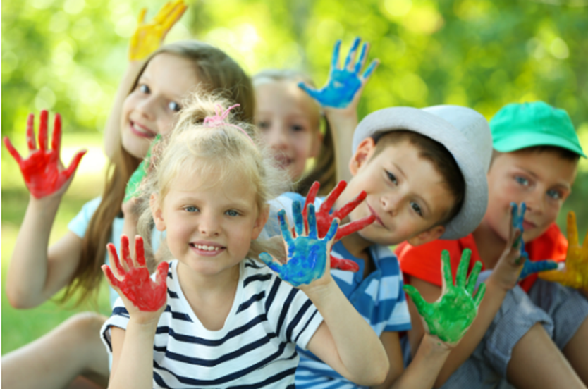 Des enfants sourient à la caméra, les mains levées et couvertes de peinture colorée, symbolisant la joie et l’expression à travers l’art.