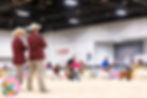An indoor Boer goat show where several people are handling Boer goats. Two livestock judges in maroon jackets and cowboy hats stand in the foreground, observing and judging the event. A colorful banner and seated spectators are visible in the background.