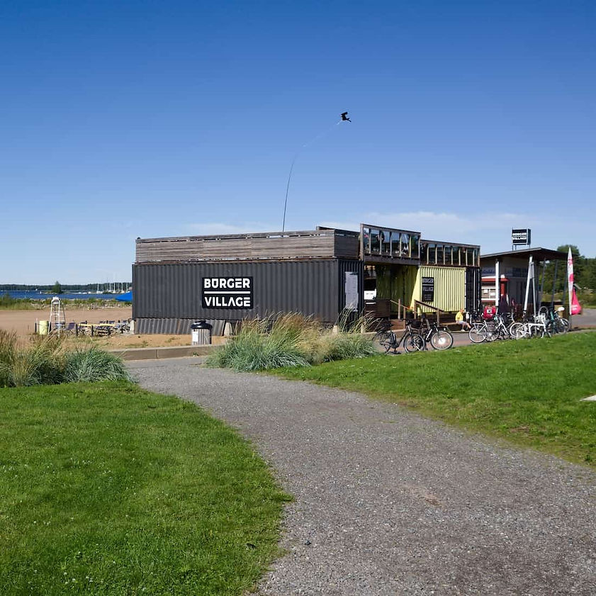 Kokkolan meripuiston hiekkarannan vieressa kuvattu Burger Village hampurilaisravintola ulkoa ja ravintolan edessa nakyy polkupyoria, vihreaa nurmikkoa ja sorapintainen kavelytie kesalla aurinkoisella saalla.