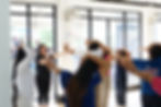 People in a room exercise with hands on heads, facing a woman leading the session. Bright windows, casual attire, and a calm atmosphere.