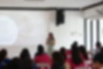 A woman presents with a microphone in a white room. Audience in pink and blue shirts faces a screen featuring text about menstrual changes.