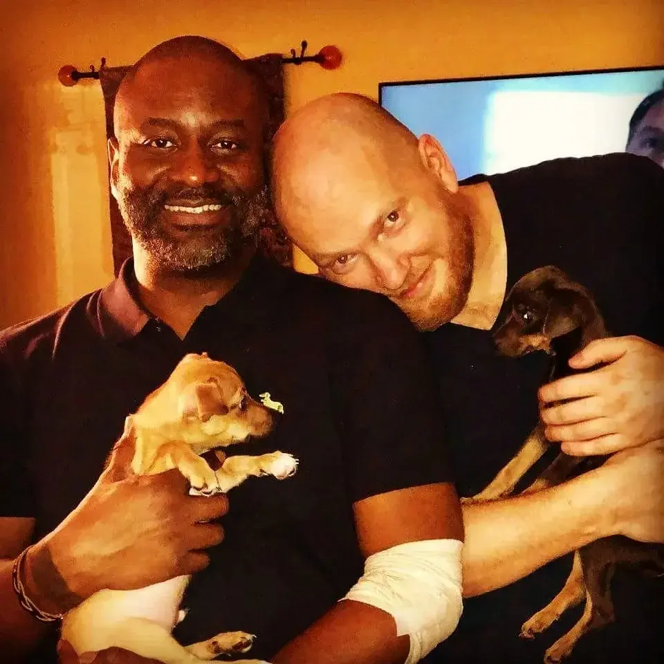 Two bearded men holding puppies with the man on the right resting his head on the other man's shoulder.