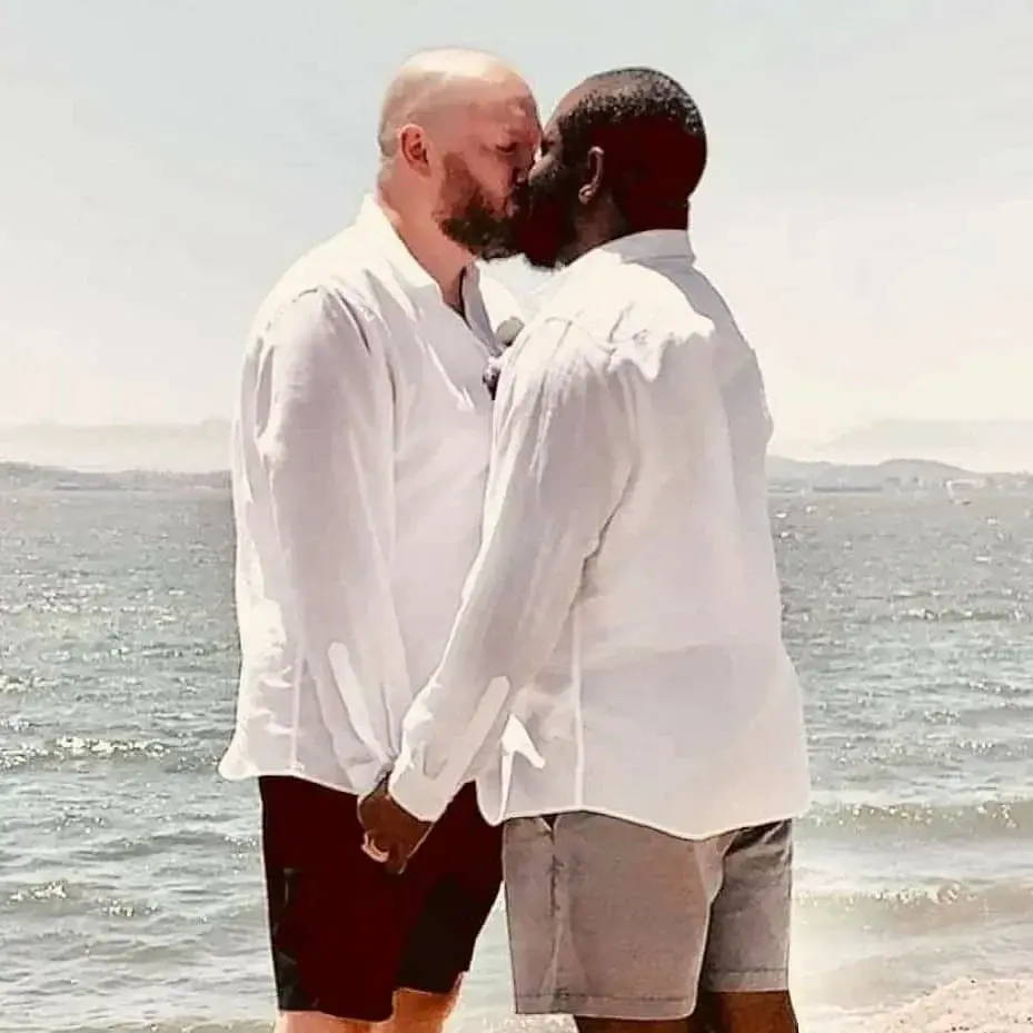 Two men wearing white, holding hands and kissing in front of the ocean waves.