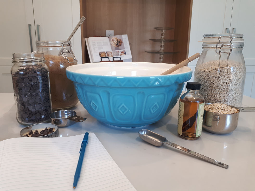 A large mixing bowl on a kitchen counter surrounded by ingredients, measuring spoons and a notebook and pen.