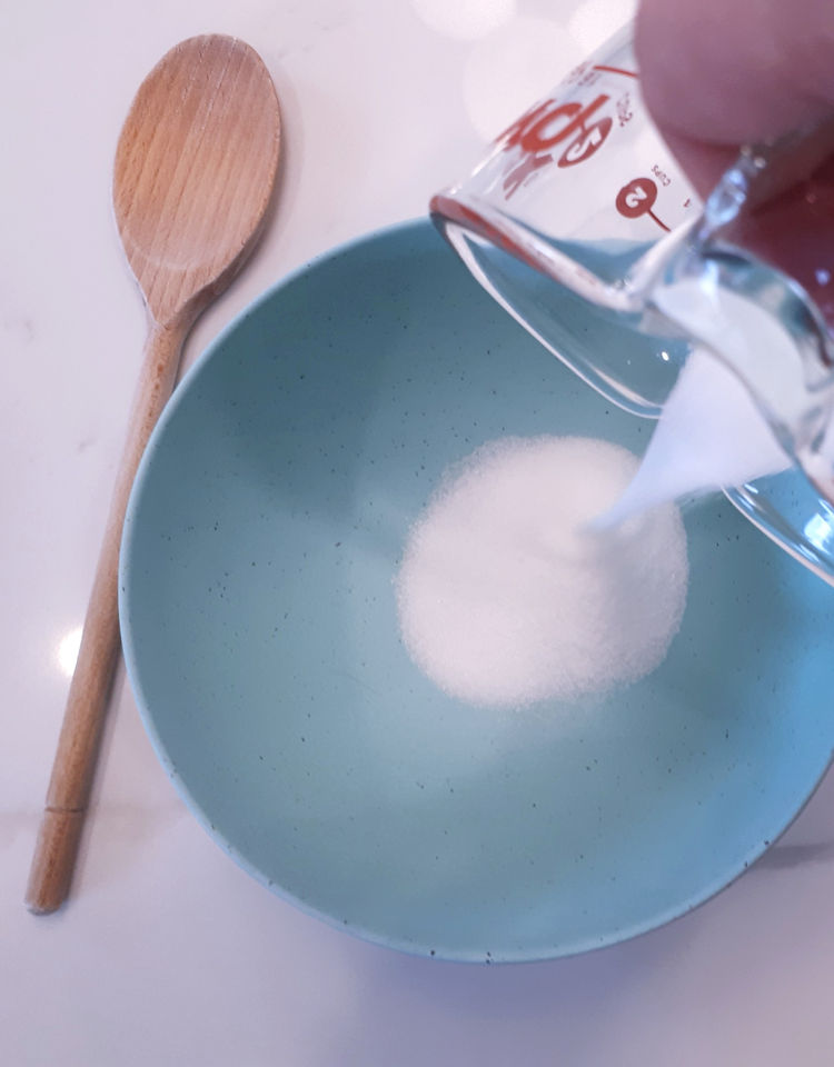 Image of sugar pouring from a glass measuring cup into a turquoise mixing bowl. A wooden spoon is on the counter beside the bowl.