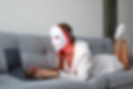 A girl with an LED mask on her face works at home on a laptop.