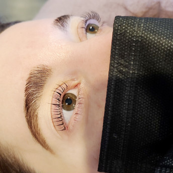 Square cropped image is a close up photo of woman's face from forehead to cheekbone. Image taken immediately after receiving eyelash lift, tinting and eyebrow lamination beauty services. Photo taken at angle to show mostly the right side of face. Over mouth and nose is a black face mask. Eyebrows are manicured, hairs look full, freshly colored eyebrow tint in brown color. Eyebrows have been brushed upwards and groomed brow design. Her eyelashes are also curly in very pretty fashion by a treatment called eyelash lifting and eyelashes are tinted to look black.