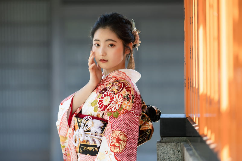 朱色の玉垣を背景に頬に手を添える振袖姿の女性