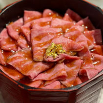 Slices of marbled pink wagyu beef drizzled with sauce on a bowl of rice, topped with gold leaf in a red bowl.