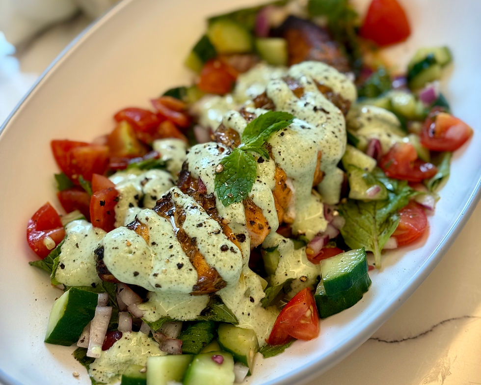 Grilled chicken salad topped with creamy green sauce, surrounded by tomatoes and cucumbers on a white plate.