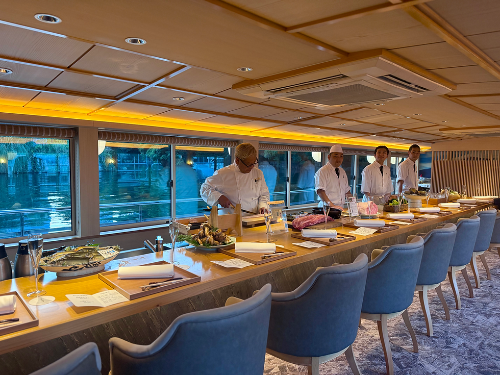 Interior of a luxury yakatabune boat featuring a long wooden counter with elegant place settings, blue upholstered chairs, and four chefs in white uniforms preparing ingredients against a backdrop of large windows overlooking the water.