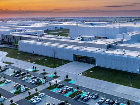 Nagy gyár naplementében, modern épületek szürke színben. Parkoló autókkal, zöld fák, narancs és kék égbolt a háttérben.