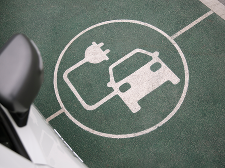 Overhead view of an electric car charging symbol painted on a green parking space, with a car partially visible in the foreground.