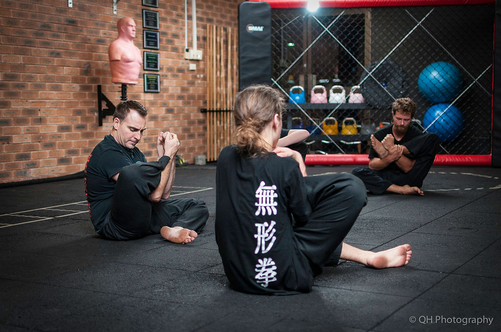 Martial arts class seated on gym mats, three people stretching in black uniforms with Chinese characters, brick wall and training gear behind.