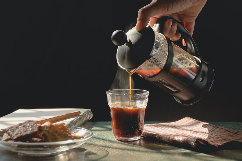Hand pours hot coffee from a French press into a glass beside pastries on a sunlit table, with a dark background and steam.