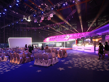 Large meeting space with lit up stage, and attendees standing around, waiting for the event to start.