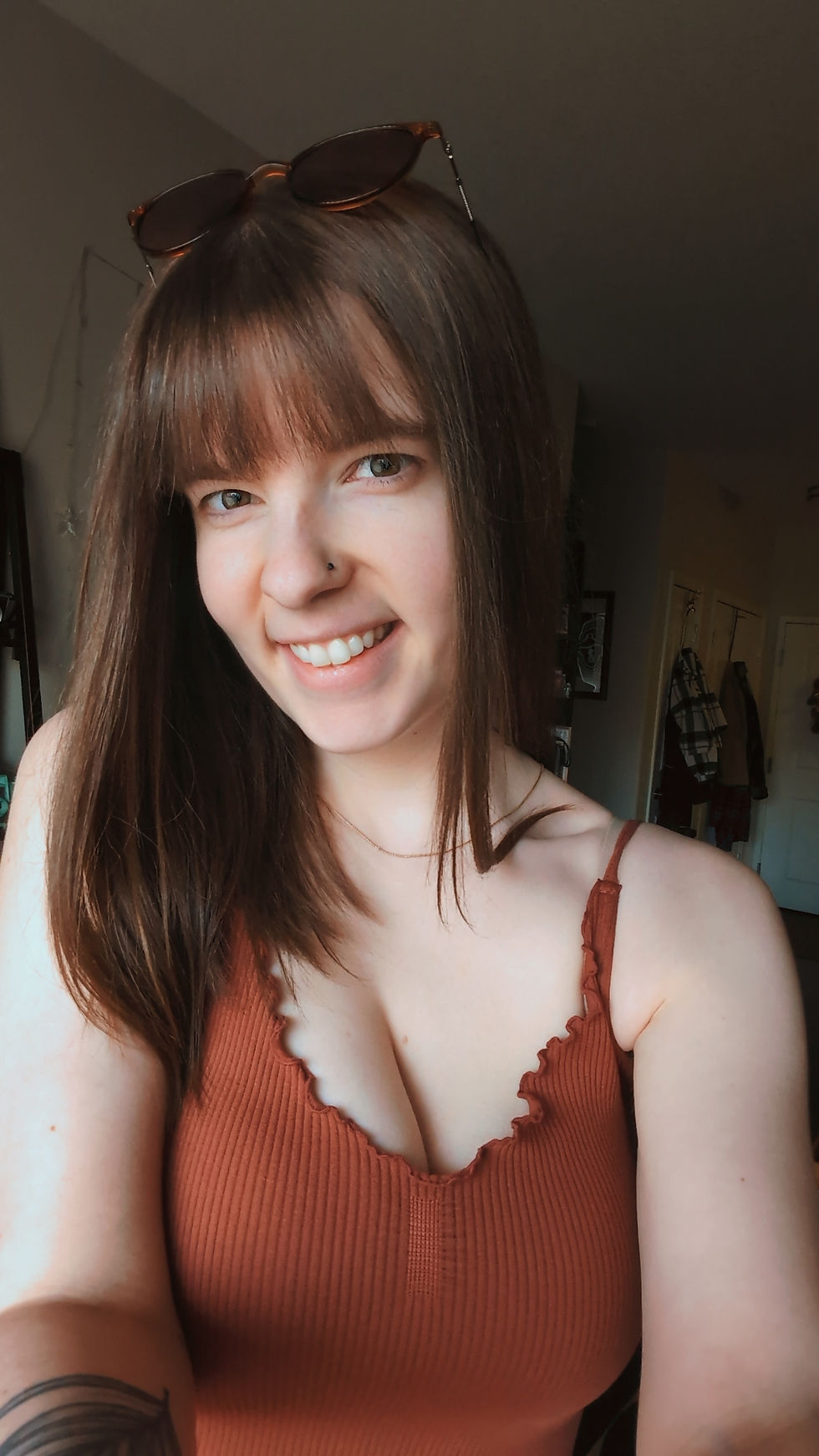 Smiling woman in a rust-colored top, wearing sunglasses on her head. Indoor setting with dim lighting. Warm, casual mood.