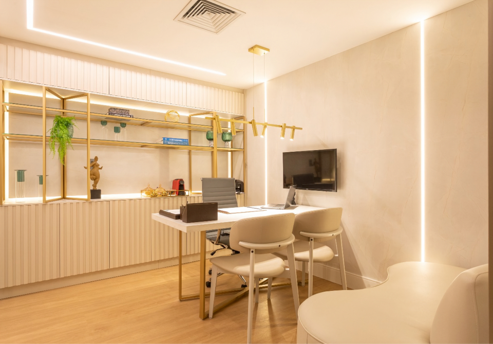 Consultório médico moderno com mesa de trabalho, cadeiras confortáveis, estante decorativa com iluminação e detalhes em dourado.







