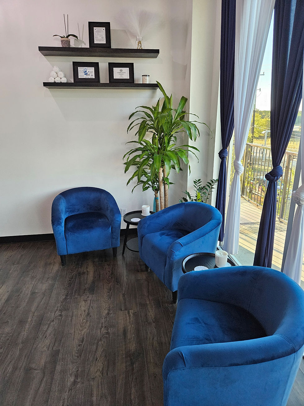 Blue velvet chairs in a cozy room with wooden floors, framed certificates on wall shelves, a potted plant, and sunlight through large windows. Luxe Thai Massage in San Antonio.