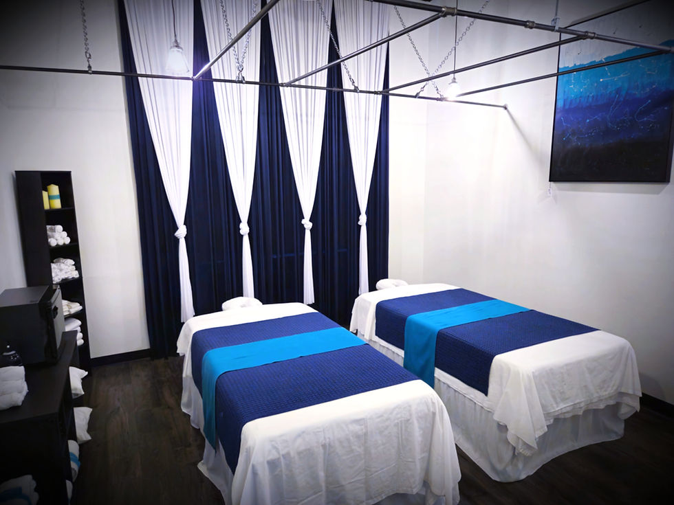 Two massage tables with blue and white linens in a spa room. White curtains hang behind, with a blue artwork on the wall, creating a serene mood. At Luxe Thai Massage in San Antonio.