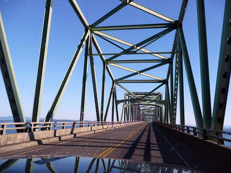 Green steel bridge seen from a car's perspective, extending over water under a clear blue sky. Prominent geometric patterns.