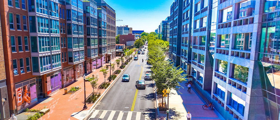 Aerial view of a city street lined with trees and modern buildings. Cars drive along the road under a clear blue sky, creating a busy yet calm urban scene.