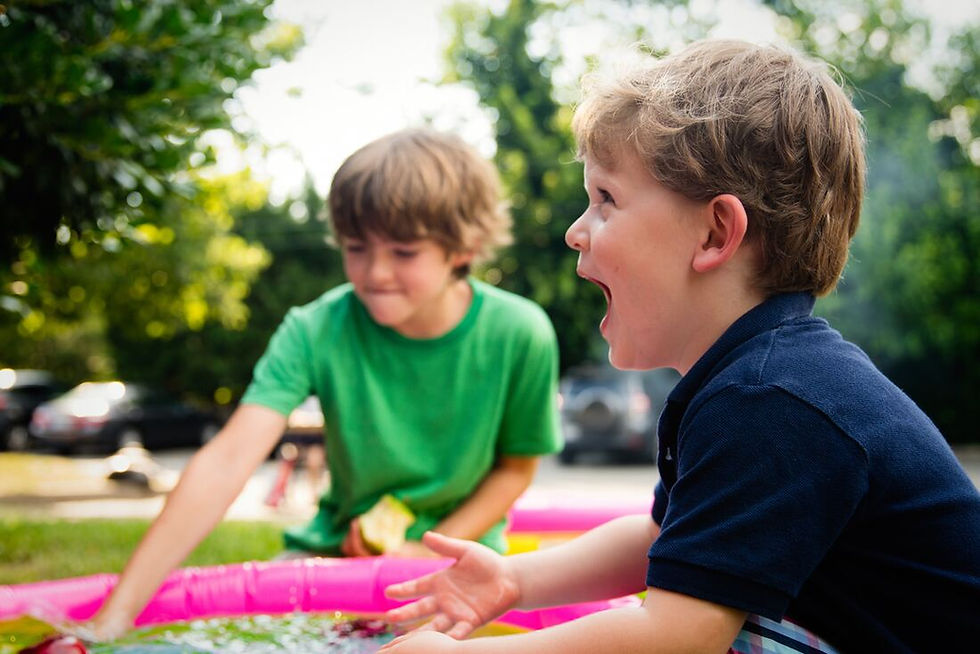 Two children play excitedly by a pink kiddie pool in a sunny garden. One wears green, the other navy. Green trees and parked cars in background.