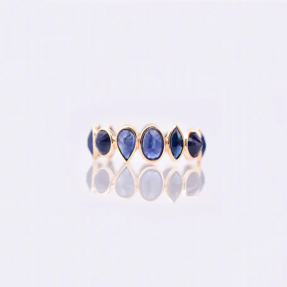 18K gold ring with marquise-shaped blue sapphire stones set in a repeating bezel pattern, photographed on a beige backdrop