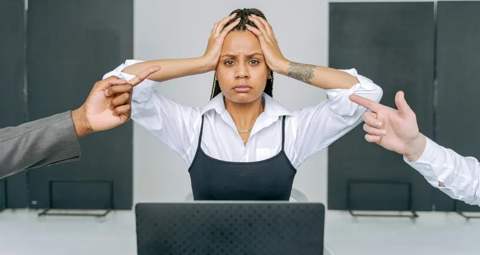 Stressed woman holding her head at a desk while colleagues point fingers at her during a tense workplace confrontation.