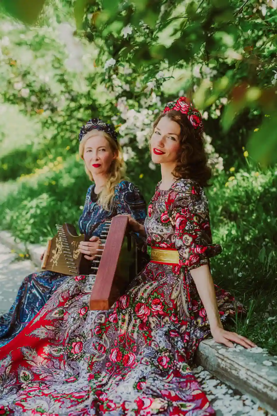 Two women in ornate, floral-patterned dresses with bright colours and headpieces, sitting outdoors and showcasing a romantic, feminine style.