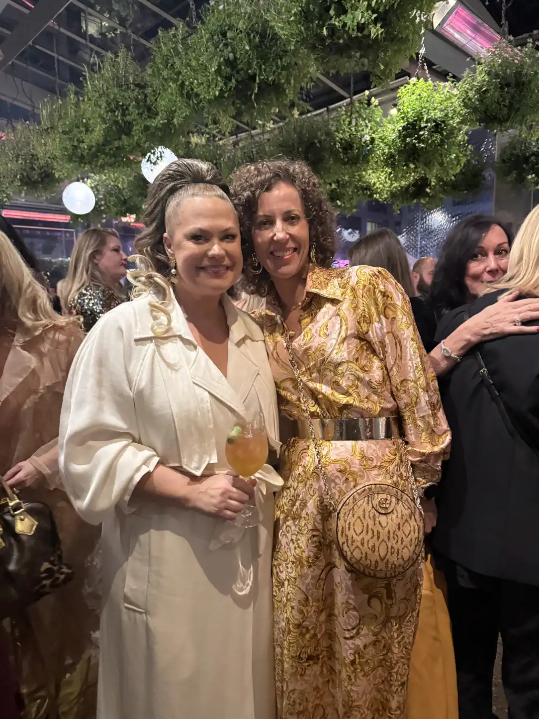 Stephanie Rumble smiles with designer Kathryn Wilson at an event, both dressed elegantly and surrounded by a lively crowd.