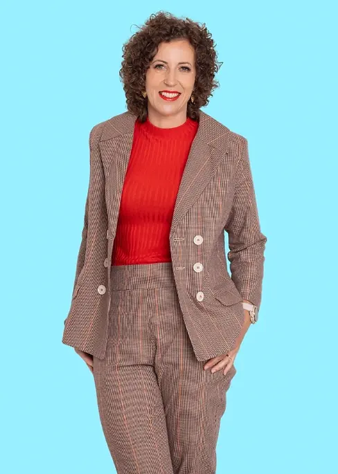 Stephanie Rumble wearing a checked brown suit with a bright red top, smiling confidently against a light blue background.