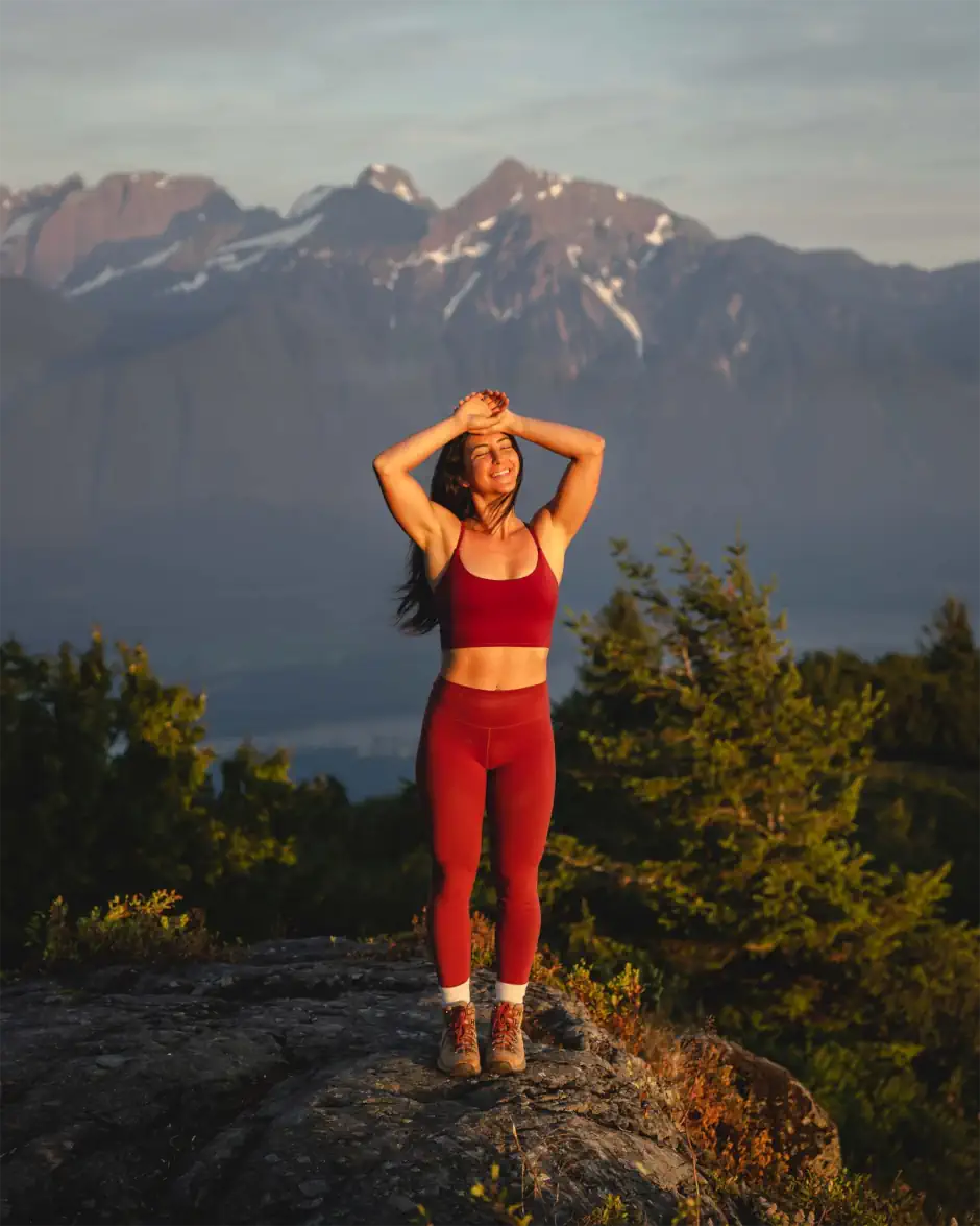 Woman standing on a rocky lookout in activewear, smiling in the sunlight with mountains and forest behind her, reflecting a natural, outdoorsy style.