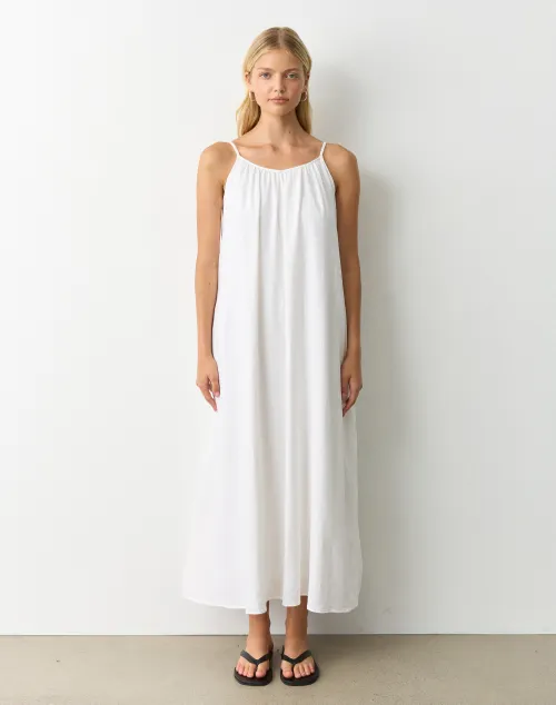 Model wearing a flowing cloud white maxi dress with thin straps, styled simply with flat sandals on a clean backdrop.