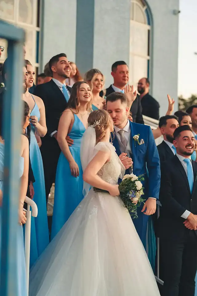 Wedding guests gathered around the bride and groom outdoors, wearing coordinated formal attire in soft blue tones.