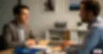Two men in a professional office setting having a serious HR conversation, seated across from each other at a desk.
