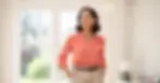 A confident woman stands indoors in soft natural light, wearing a coral blouse and beige trousers, looking thoughtfully into the distance. The scene feels calm, professional, and empowering.