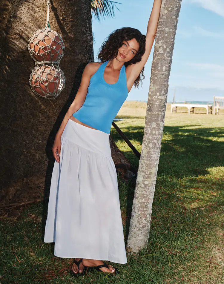 Model wearing a flowing white long skirt styled with a fitted blue tank top in a relaxed outdoor summer setting.