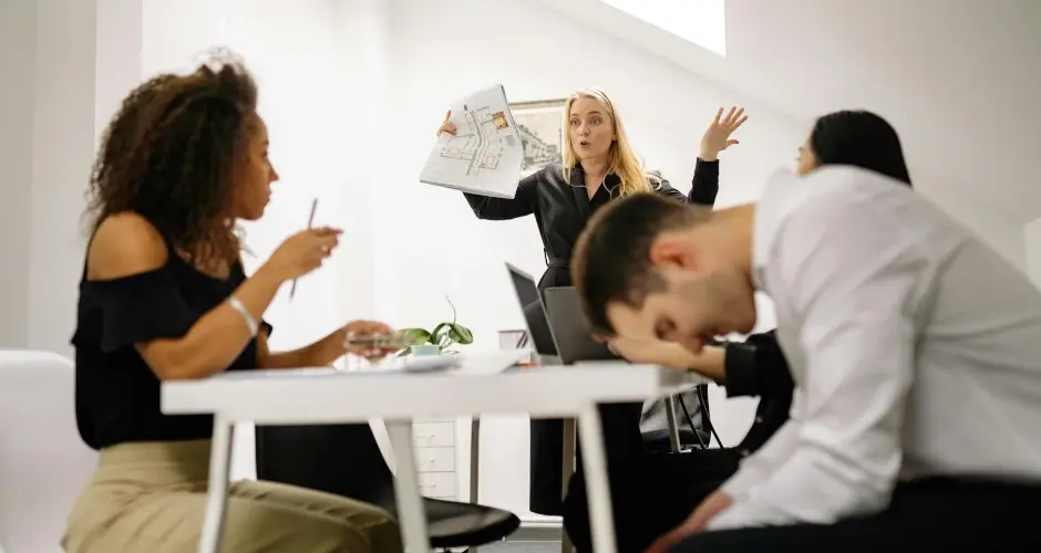 Tense office meeting where a woman speaks assertively while others appear frustrated, disengaged, or overwhelmed.