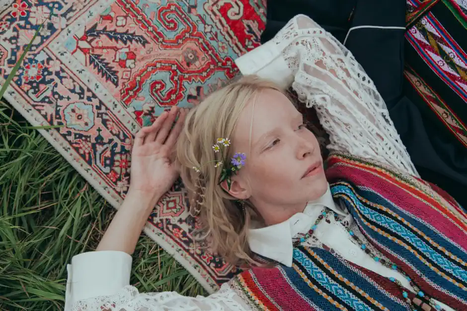 Woman lying on a patterned rug wearing colourful, embroidered clothing with lace details and flowers in her hair, expressing a creative, bohemian style.